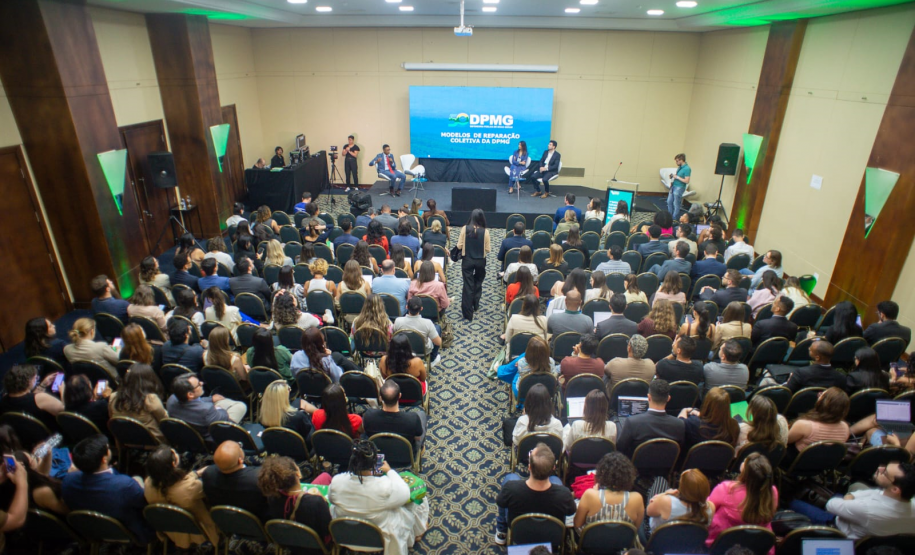 Vista superior do fundo de um auditório lotado, com a plateia voltada para um palco com iluminação azul. No telão, lê-se "Modelos de Reparação Coletiva da DPMG", enquanto palestrantes debatem sentados no palco.