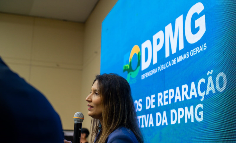 Uma mulher de perfil, com cabelos longos, fala ao microfone. Atrás dela, um painel de LED azul exibe o logotipo da DPMG e o texto parcial "Modelos de Reparação Coletiva".