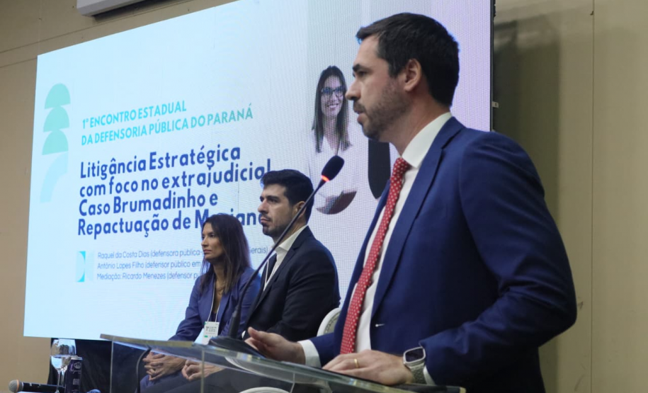 Um homem de terno azul e gravata vermelha discursa em um púlpito. Atrás dele, duas pessoas estão sentadas no palco, e um telão exibe o título da palestra: "Litigância Estratégica com foco no extrajudicial".