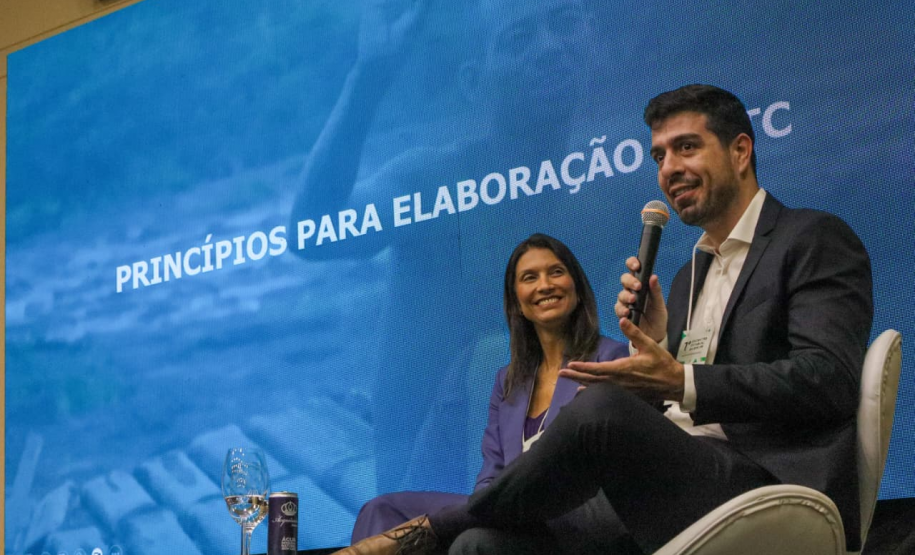 Um homem de terno escuro e camisa branca fala ao microfone, sentado em uma poltrona branca no palco. Ao seu lado, uma mulher de blazer azul sorri enquanto observa a plateia. Atrás deles, um grande painel de LED azul exibe a frase "PRINCÍPIOS PARA ELABORAÇÃO".