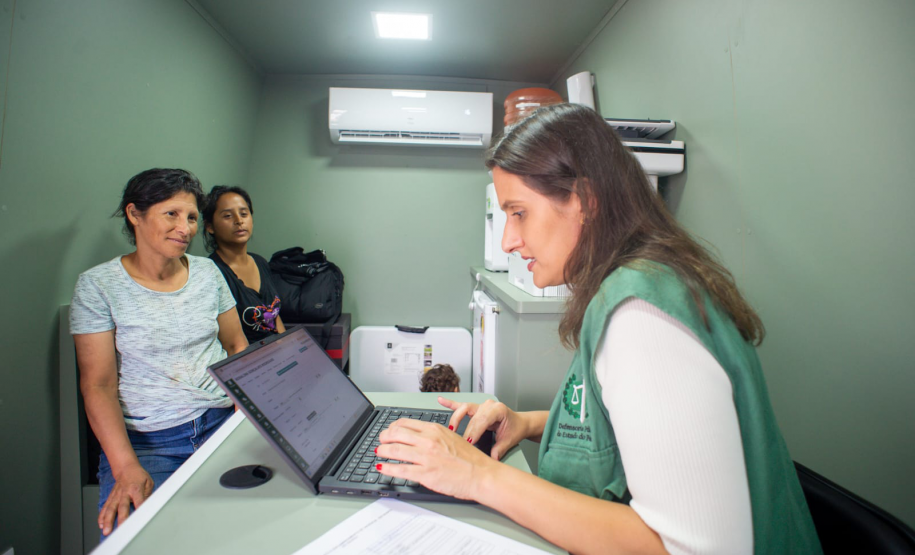 Defensora Mariana, com colete verde da Defensoria, atende duas mulheres, uma com blusa azul claro e outra com camiseta escura. Elas estão no interior da van dos direitos. Ao fundo um ar condicionado e uma mesinha com impressora.