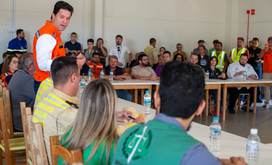 Em uma sala de reunião, o secretário das cidades do Estado, Guto Silva, está em pé vestindo colete laranja (Defesa Civil/Casa Militar), fala com o público sentado nas mesas brancas. Em primeiro plano, uma mulher e um homem de colete verde da Defensoria Pública ouvem.