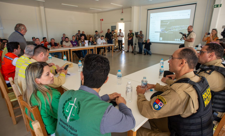 Cena de reunião com várias pessoas sentadas em mesas brancas em "U" em uma sala iluminada. Em primeiro plano, defensores públicos juntos de oficiais da Polícia Militar e outras autoridades, sentados. Há uma tela de projeção exibindo um mapa ao fundo.