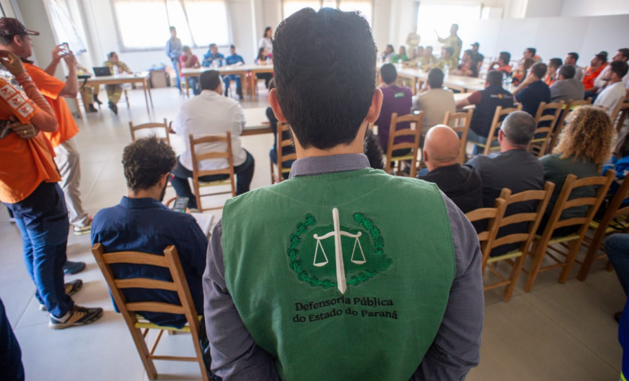 Cena de reunião com várias pessoas sentadas em mesas brancas em "U" em uma sala iluminada. Em primeiro plano, defensores públicos juntos de oficiais da Polícia Militar e outras autoridades, sentados. Há uma tela de projeção exibindo um mapa ao fundo.