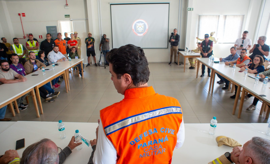 Em uma sala de reunião, o secretário das cidades do Estado, Guto Silva, está em pé vestindo colete laranja (Defesa Civil/Casa Militar), fala com o público sentado nas mesas brancas.