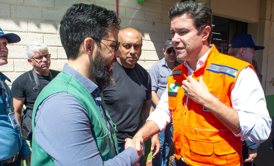 Dois homens brancos se cumprimentam com um aperto de mãos. O da esquerda, o defensor público Fernando Redede, com barba e óculos, veste um colete verde da defensoria. À direita, o secretário das cidades do Estado, Guto Silva, veste um colete de segurança laranja, com a mão esquerda sobre o peito. Há outras pessoas ao fundo.