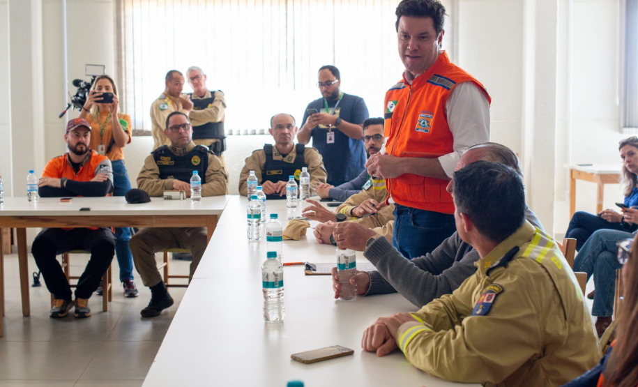 Em uma sala de reunião, o secretário das cidades do Estado, Guto Silva, está em pé vestindo colete laranja (Defesa Civil/Casa Militar), fala com o público sentado nas mesas brancas.