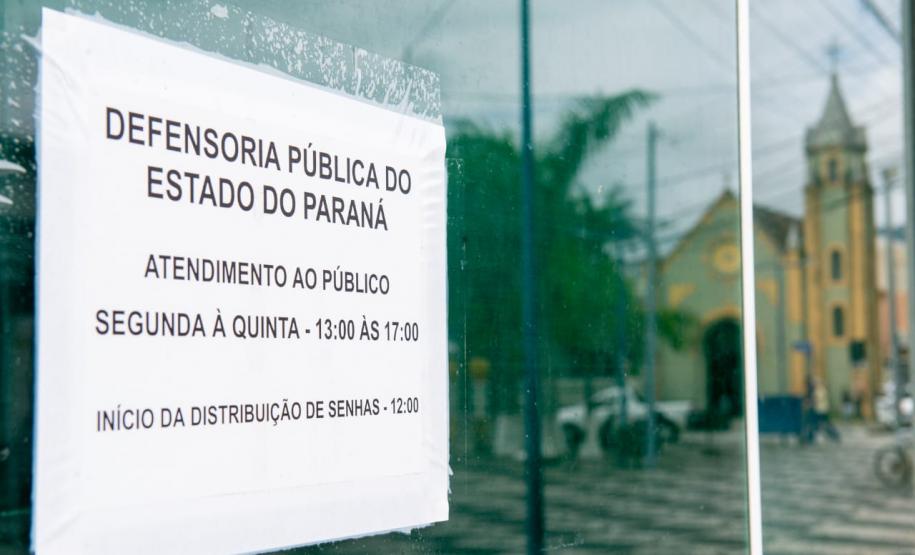 Imagem da placa da Defensoria com reflexo na porta de vidro da Câmara em que se vê o reflexo da igreja na praça. da igreja da praça em Matinhos.