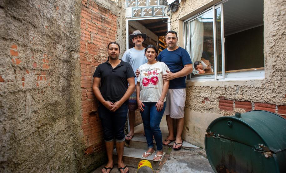 Família com quatro pessoas adultas posa em frente à sua casa. Muros e paredes sem reboco compõe o cenário.