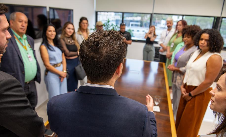 Marcel Jeronymo Lima Oliveira aparece de costas e ao fundo as autoridades e lideranças de movimentos sociais durante a posse como ouvidor-geral da Defensoria Pública do Estado do Paraná.