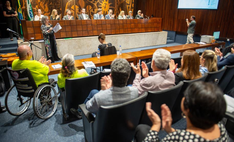 Cadeirante em primeiro plano e público ao redor aplaudem lançamento do NUPED na ALEP.