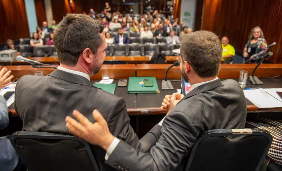 Defensores públicos de costas se cumprimentam na mesa diretora do lançamento do NUPED. Ao fundo o público presente.