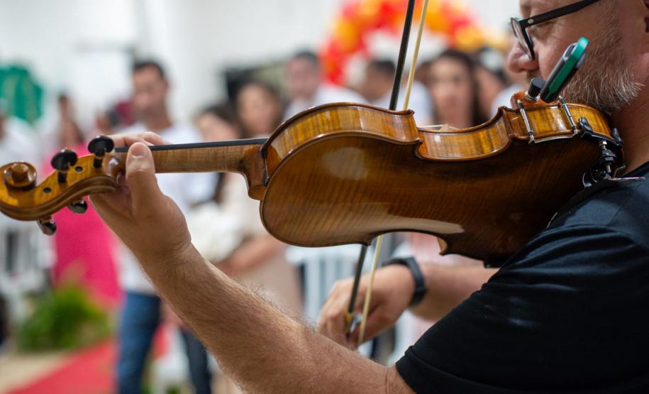 Imagem mostra um violino em primeiro plano e os noivos e noivas ao fundo desfocados.