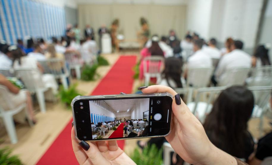 Imagem mostra um celular em primeiro plano e ao fundo a cerimônia com 20 casais.