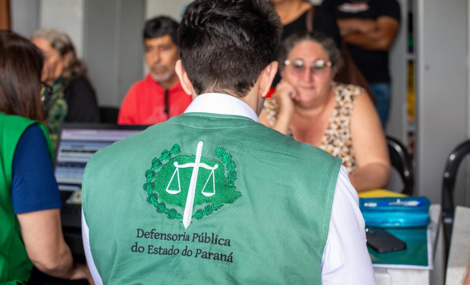 Imagem mostra Defensor Público de costas com colete da instituição realizando atendimento de uma senhora em mutirão no Litoral do Paraná.
