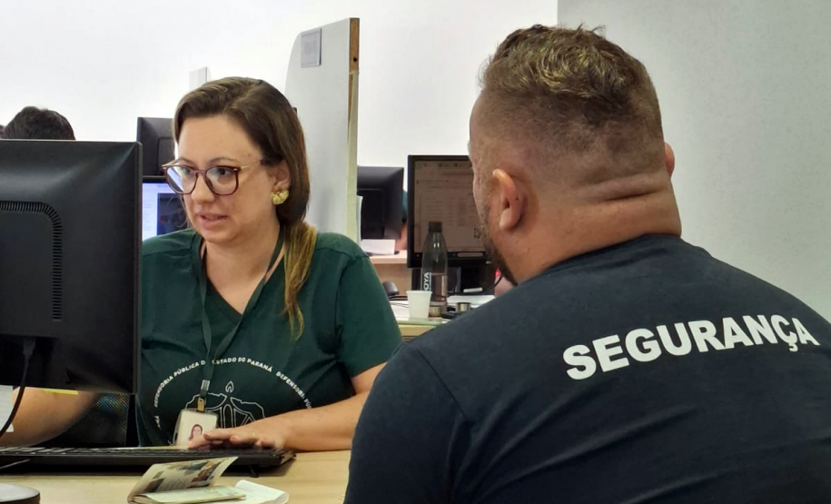 Usuário de costas utilizando uma camiseta com a palavra "segurança" é atendido por uma servidora da Defensoria no Mutirão realizado em Maringá.