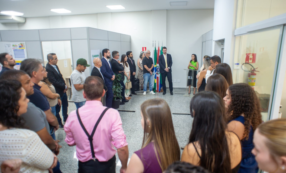 Uma mulher de blusa verde fala ao microfone em um pequeno púlpito, de frente para uma plateia que a assiste de pé em uma sala ampla. Atrás dela, junto a bandeiras oficiais, estão outras autoridades.