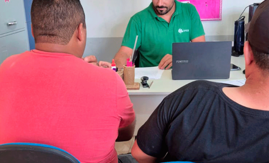 Um close do atendimento onde o funcionário de camisa verde consulta documentos e um notebook. De frente para ele, dois homens aguardam sentados de costas para a câmera em cadeiras de assento azul.