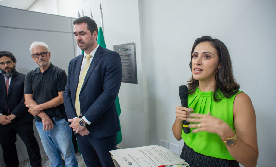 Em um plano mais aproximado, a mulher de blusa verde discursa ao microfone com expressão atenta, apoiada em um púlpito com papéis. Logo atrás dela, três homens aguardam em pé, escutando-a com seriedade.