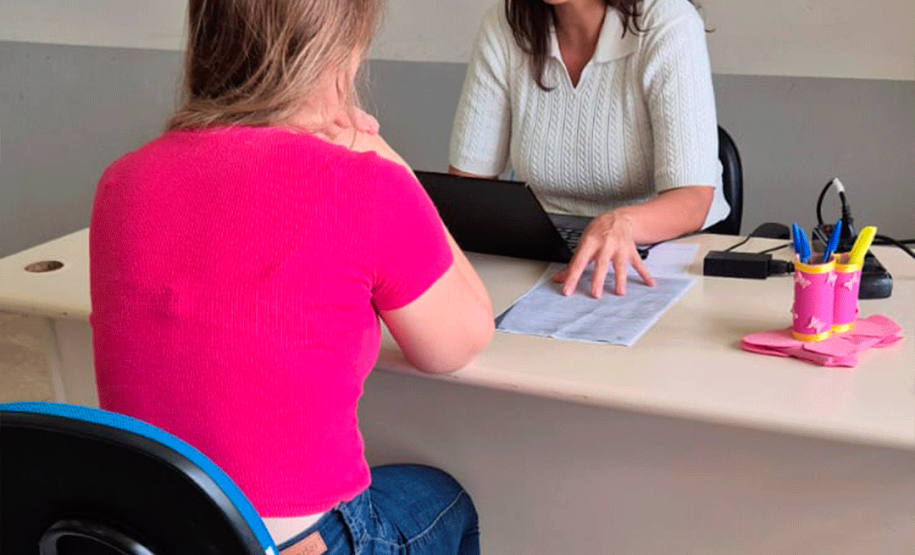 Uma mulher de blusa branca atende uma cidadã sentada à sua frente, que veste blusa rosa e calça jeans e é vista de costas. A atendente fala enquanto aponta para papéis sobre a mesa, ao lado de um notebook.
