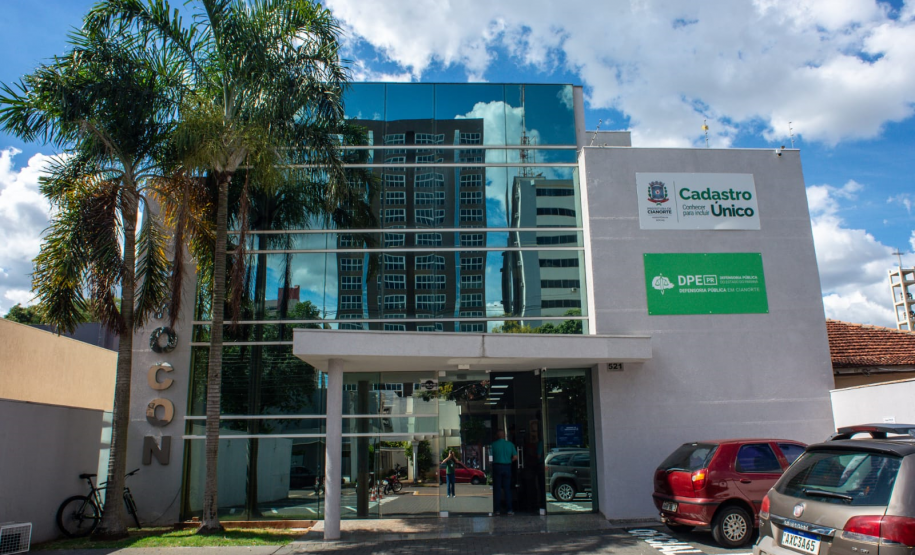 Fachada de um prédio moderno com grande painel de vidro espelhado. À direita, na parede branca, destacam-se placas do "Cadastro Único" e da "DPE-PR em Cianorte". Há palmeiras e carros estacionados na entrada.