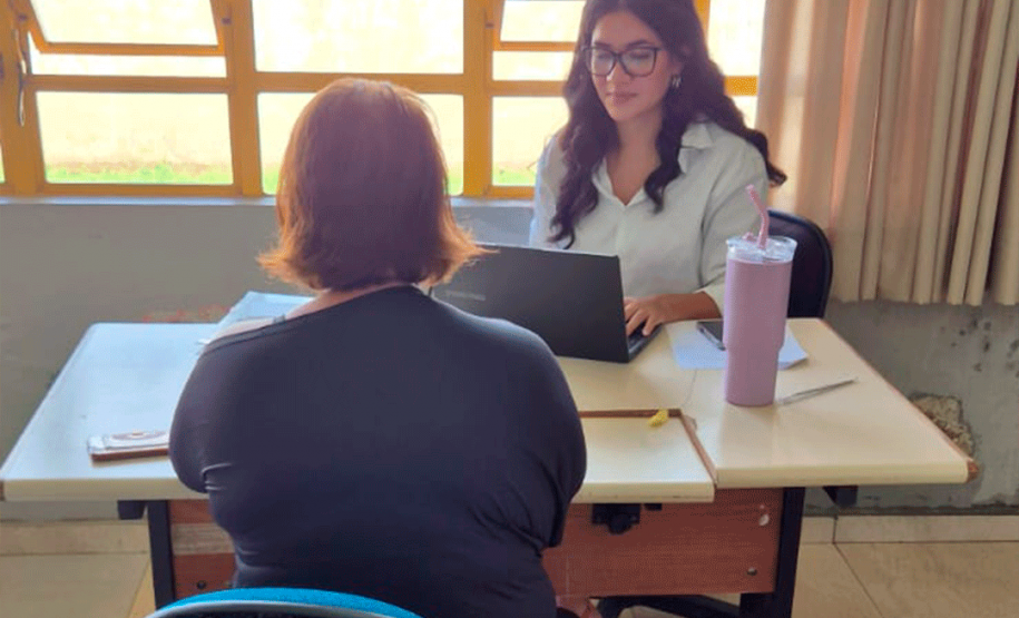 Próximo a uma grande janela com esquadrias amarelas, uma mulher de óculos e camisa branca digita em um notebook. Sentada de costas em uma cadeira azul, uma mulher de roupa escura recebe o atendimento.