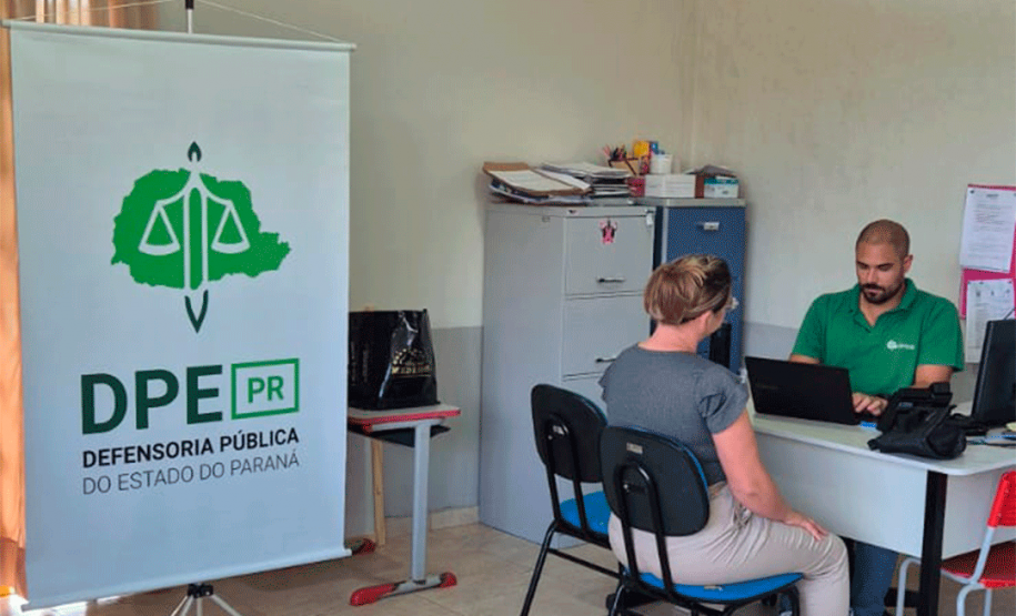 Visão mais ampla da sala com um banner vertical da DPE-PR em destaque à esquerda. Ao fundo, o funcionário de verde usa o notebook enquanto atende uma mulher de blusa cinza sentada à sua frente.