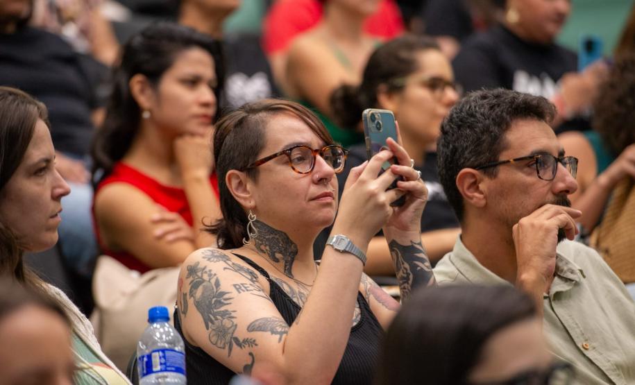 Imagem mostra detalhe do público presente. Em primeiro plano uma mulher com tatuagens registra o evento com celular.