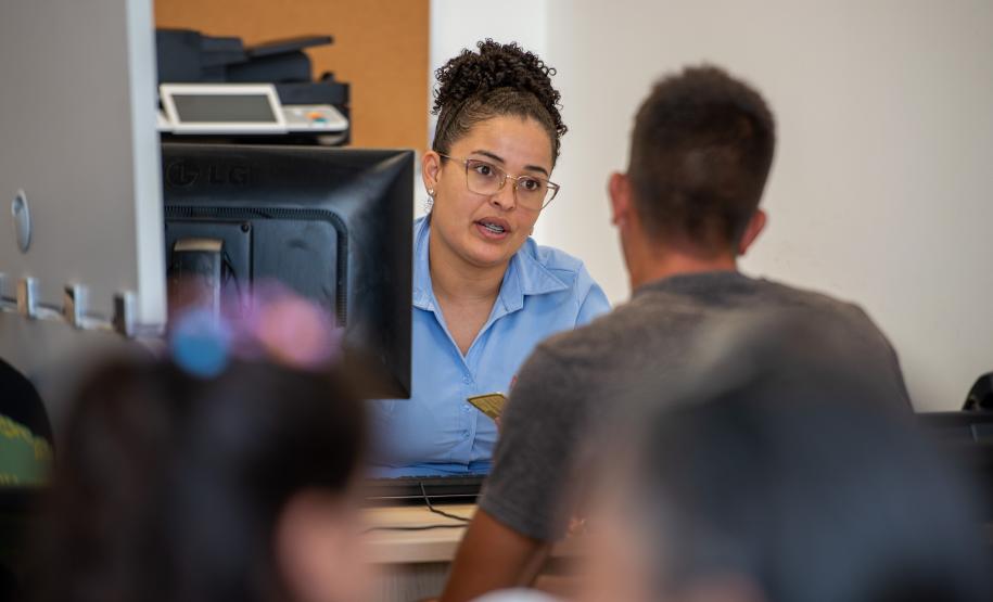 Imagem mostra uma servidora da DPE-PR realizando atendimento a um cidadão. Ele aparece de costas. Em primeiro plano desfocado aparecem cabeças de pessoas na fila de espera.