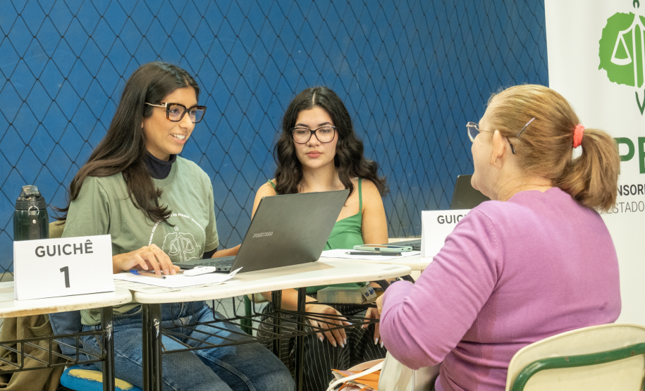 Imagem mostra uma mulher com cabelos presos de costas recebendo atendimento de duas servidoras da Defensoria Pública do Paraná.