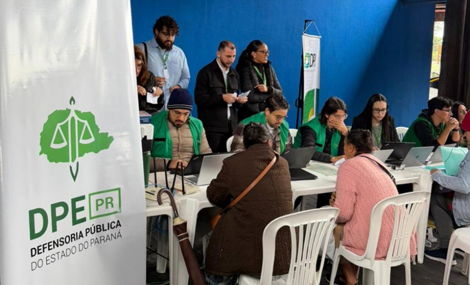 Em um espaço com parede azul, profissionais com coletes verdes usam notebooks em mesas brancas para atender cidadãos sentados à sua frente. À esquerda, destaca-se um banner vertical da Defensoria Pública do Paraná.