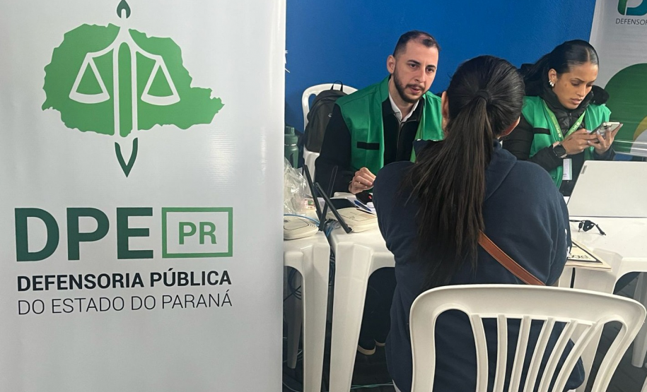 Close-up de um atendimento onde um servidor de colete verde conversa com uma mulher sentada de costas. Ao lado dele, outra profissional manuseia o celular, com um banner da DPE-PR em grande destaque no lado esquerdo.