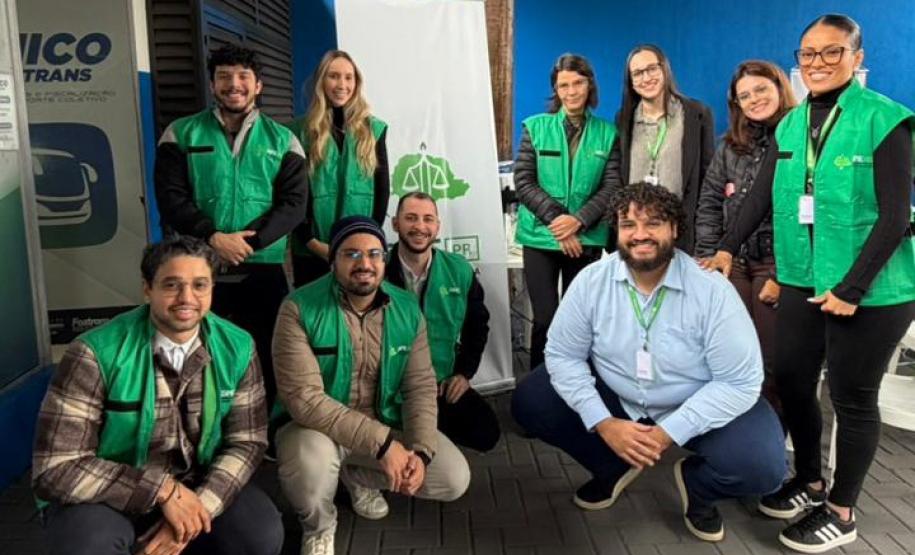 Dez profissionais da equipe da DPE-PR posam sorridentes para uma foto em grupo. A maioria veste coletes verdes da instituição, dividindo-se entre pessoas em pé e agachadas ao redor de um banner institucional.