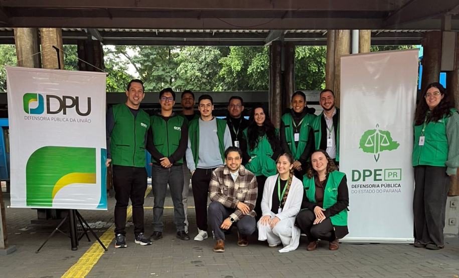 Uma equipe de doze pessoas posa para foto em um espaço público com piso tátil amarelo no chão. A equipe, com vários membros usando coletes verdes, está posicionada entre os banners da Defensoria Pública da União (DPU) e da DPE-PR.