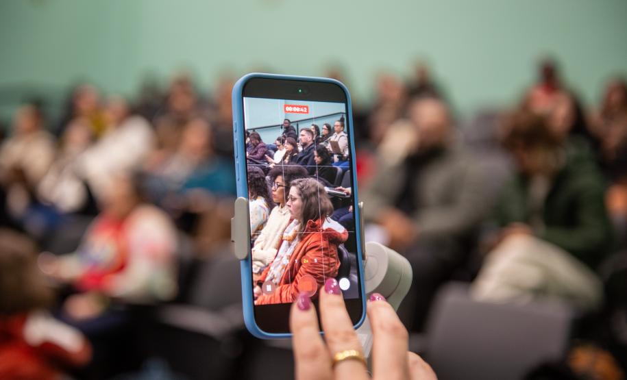 Imagem mostra close em celular mostrando o auditório cheio ao fundo.