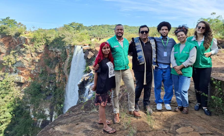 Equipe da Defensoria posa ao lado de indígenas e ao fundo uma cachoeira que fica dentro do território.