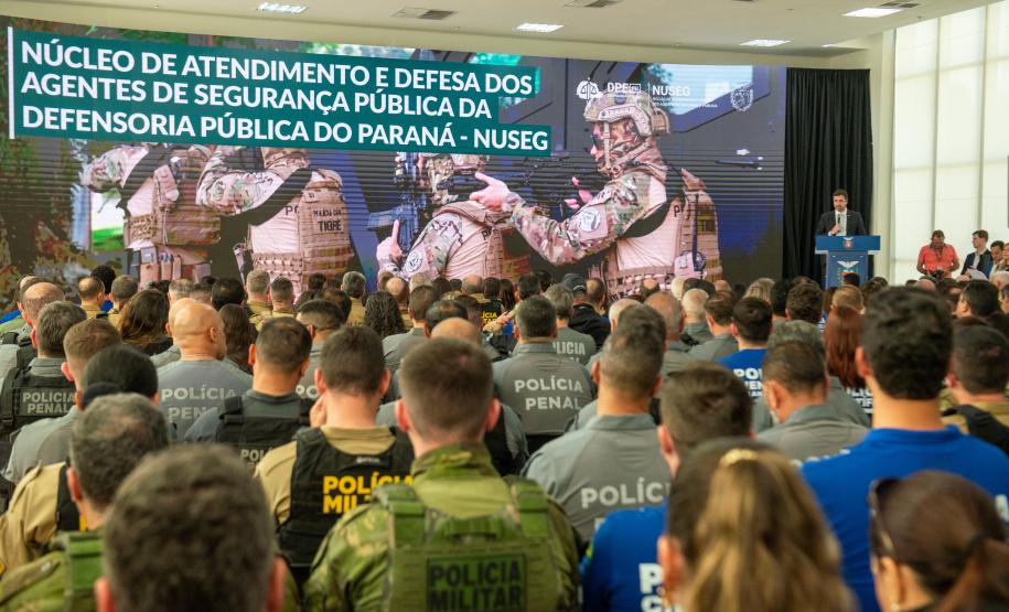 Imagem mostra o salão de atos do Palácio Iguaçu lotado com oficiais das forças de segurança. Ao fundo letreiro sobre o lançamento do NUSEG e o DPG Matheus Munhoz discursando.