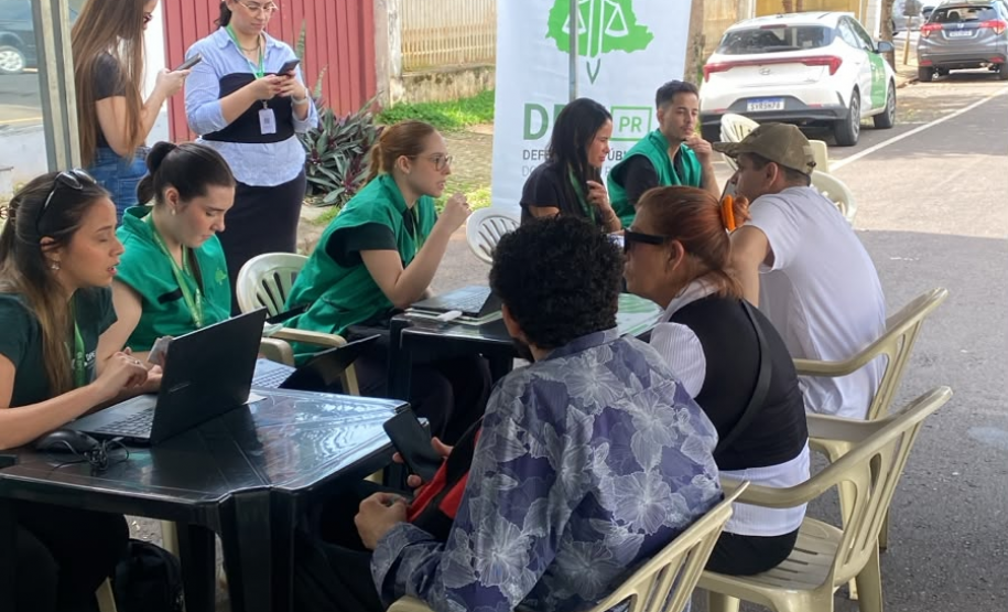 Em uma tenda de atendimento na rua a equipe da Defensoria Pública conversa e orienta as pessoas sentadas à frente das mesas. Ao fundo, destaca-se um banner branco e verde da DPE-PR.
