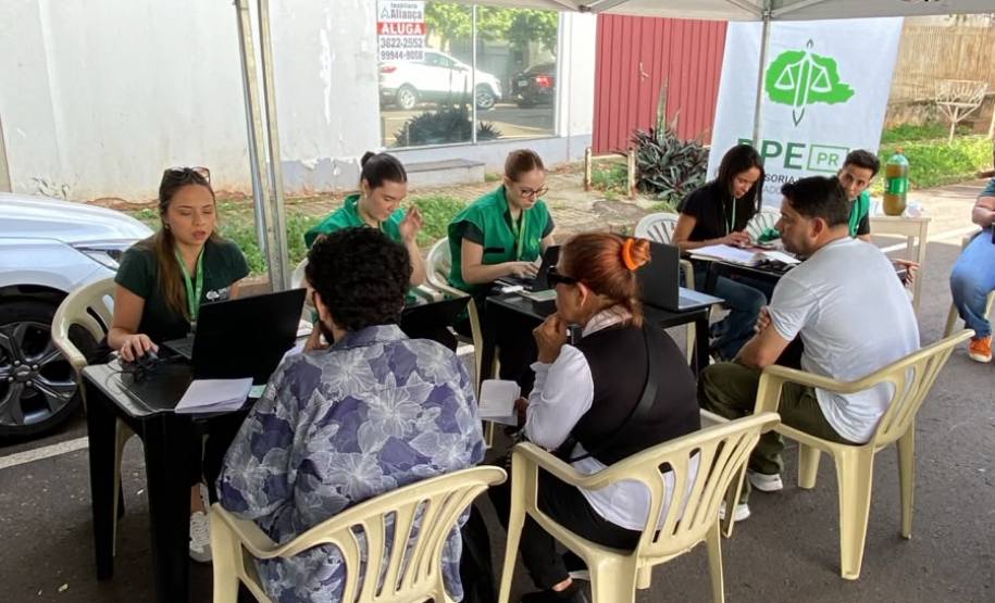 Atendimento ao ar livre sob uma tenda branca. Funcionárias de colete verde da DPE-PR utilizam notebooks em mesas para atender cidadãos que estão sentados em cadeiras de plástico branco.