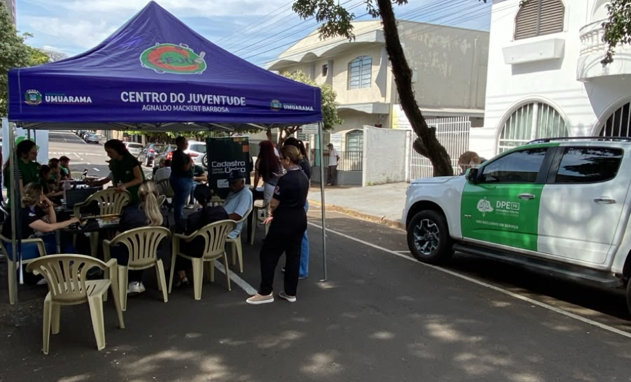 Uma tenda azul com a inscrição "Centro da Juventude" montada na rua abriga pessoas em atendimento. Estacionada logo ao lado, sob a sombra de uma árvore, está uma caminhonete branca com a logo verde da DPE-PR.
