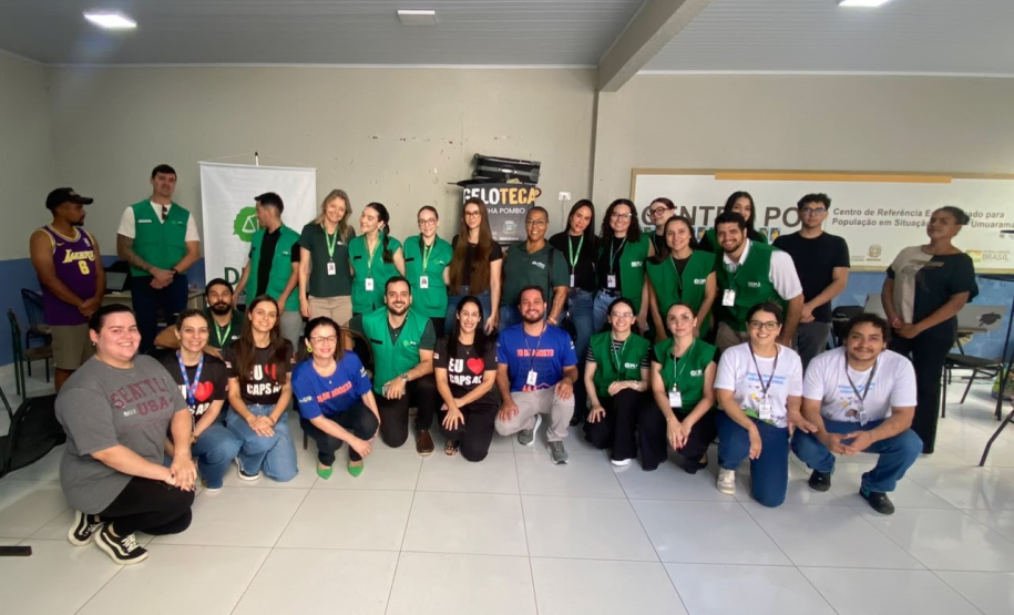 Fotografia de um grande grupo posando sorridente em um salão claro. Muitas pessoas vestem o colete verde da DPE-PR, acompanhadas por outras em trajes casuais. Ao fundo, há letreiros indicando "Centro POP".