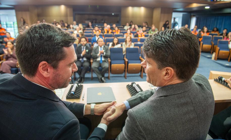DPG Matheus Munhoz e presidente do TCE apertam as mãos, ao fundo a platéia.