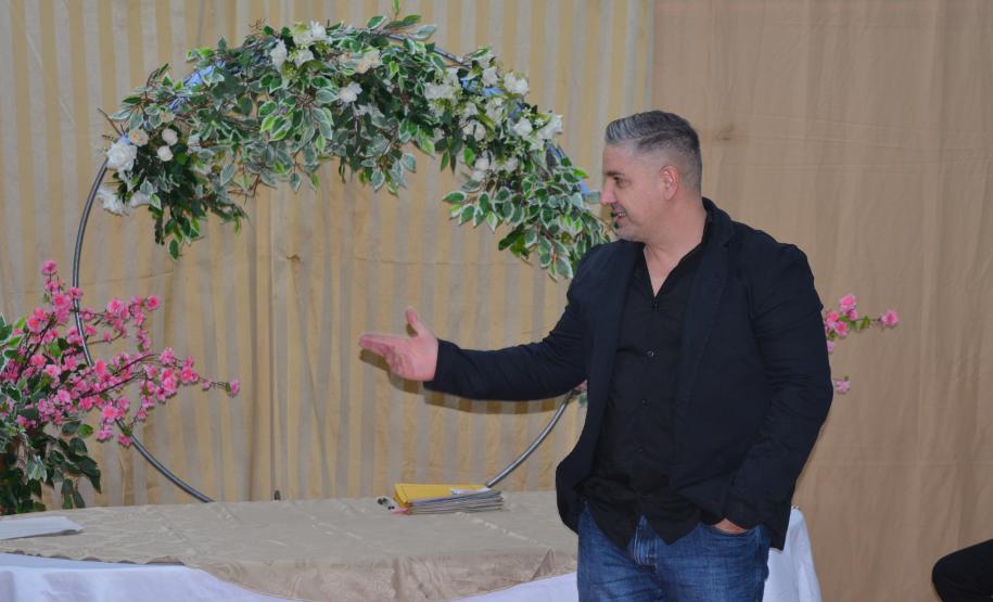 Um homem de blazer escuro, camisa preta e calça jeans gesticula com a mão direita. Ele está posicionado em frente a um arco decorativo adornado com flores brancas e folhagens, ladeado por arranjos de flores rosas.