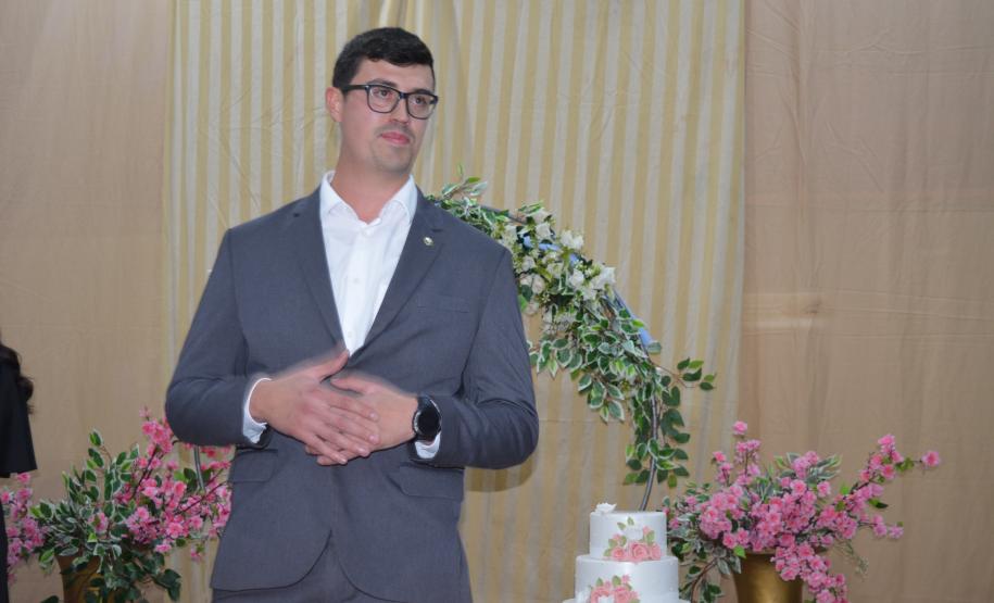 Um homem de terno cinza, camisa branca e óculos está de pé com as mãos cruzadas à frente, olhando levemente para o lado. Próximo a ele há um bolo branco de três andares enfeitado com flores rosas, com o arco floral ao fundo.