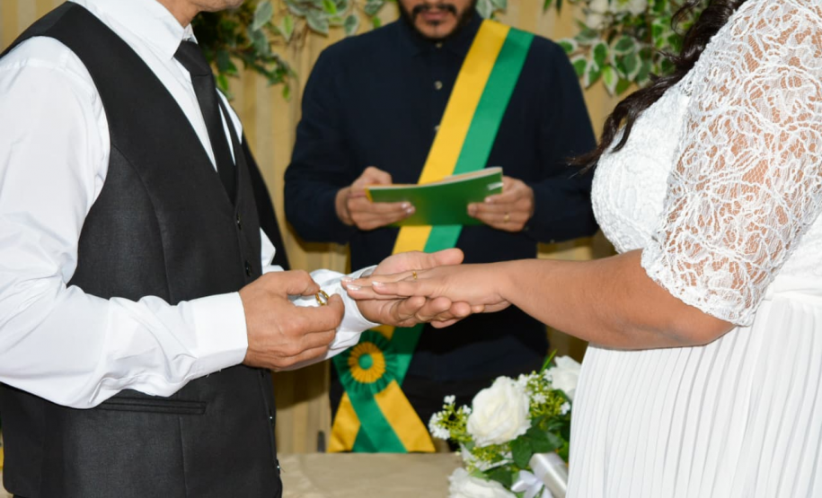 Detalhe do momento exato da troca de alianças. O noivo, de colete preto e camisa branca, coloca a aliança no dedo da noiva de vestido rendado, enquanto o celebrante com faixa verde e amarela observa desfocado ao fundo.