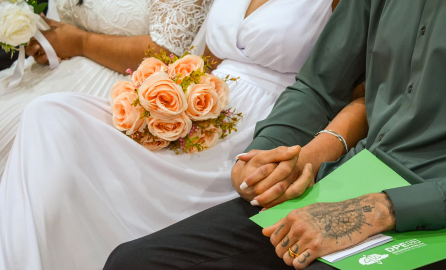 Close no colo de um casal sentado. A noiva, de vestido branco, segura um buquê de rosas pêssego, enquanto o companheiro ao lado apoia as mãos, uma delas tatuada e com aliança, sobre uma pasta verde da DPE-PR.