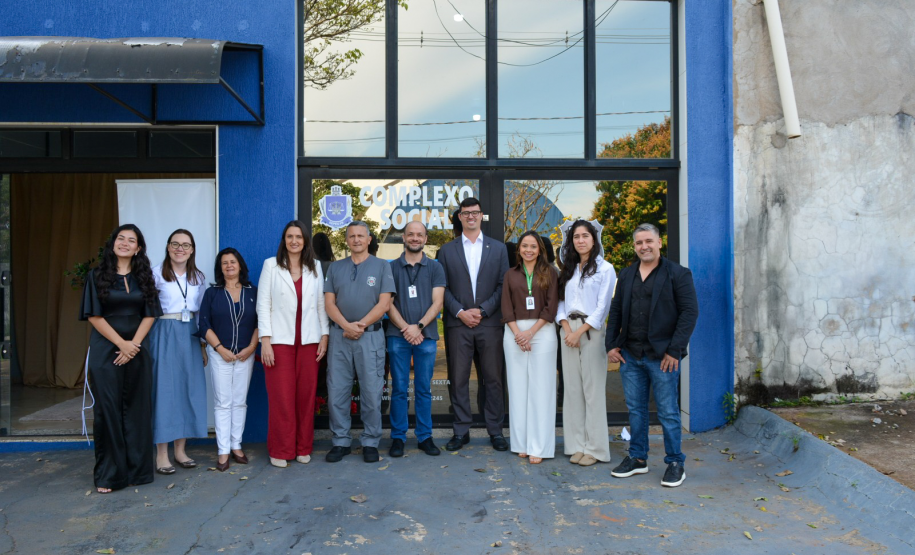 Um grupo de dez pessoas posa sorridente para uma fotografia em frente à fachada azul e às portas de vidro de um prédio identificado como "COMPLEXO SOCIAL". A equipe veste trajes sociais casuais, e alguns usam crachás de identificação no pescoço.