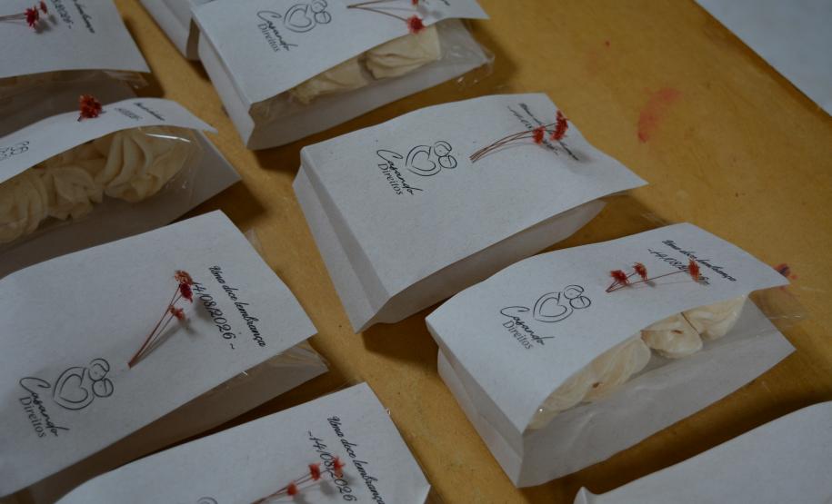 Foco em diversas lembrancinhas de papel branco dispostas sobre uma mesa amarela. As embalagens contêm suspiros, trazem a logo "Casando Direitos" e a data "14/08/2026", e são enfeitadas com pequenas flores vermelhas secas.