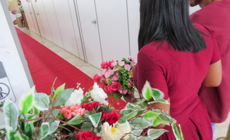 Um casal, com a mulher vestindo um vestido vermelho, caminha por um corredor coberto por um tapete vermelho. Em primeiro plano e ligeiramente desfocados, arranjos de flores brancas, rosas e vermelhas enfeitam a lateral do caminho.