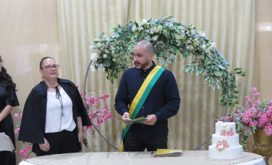 Um celebrante com uma faixa verde e amarela no peito está posicionado atrás de uma mesa com um pequeno bolo de casamento enfeitado com flores. Ao lado dele, uma mulher de blusa preta e branca acompanha a cerimônia em frente a um arco floral.